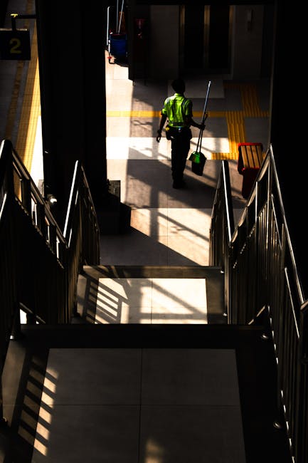 A professional cleaning technician wearing a white protective suit, face mask, and orange gloves is operating a carpet or surface cleaning machine in a modern, well-lit room with grey walls, tall wind
